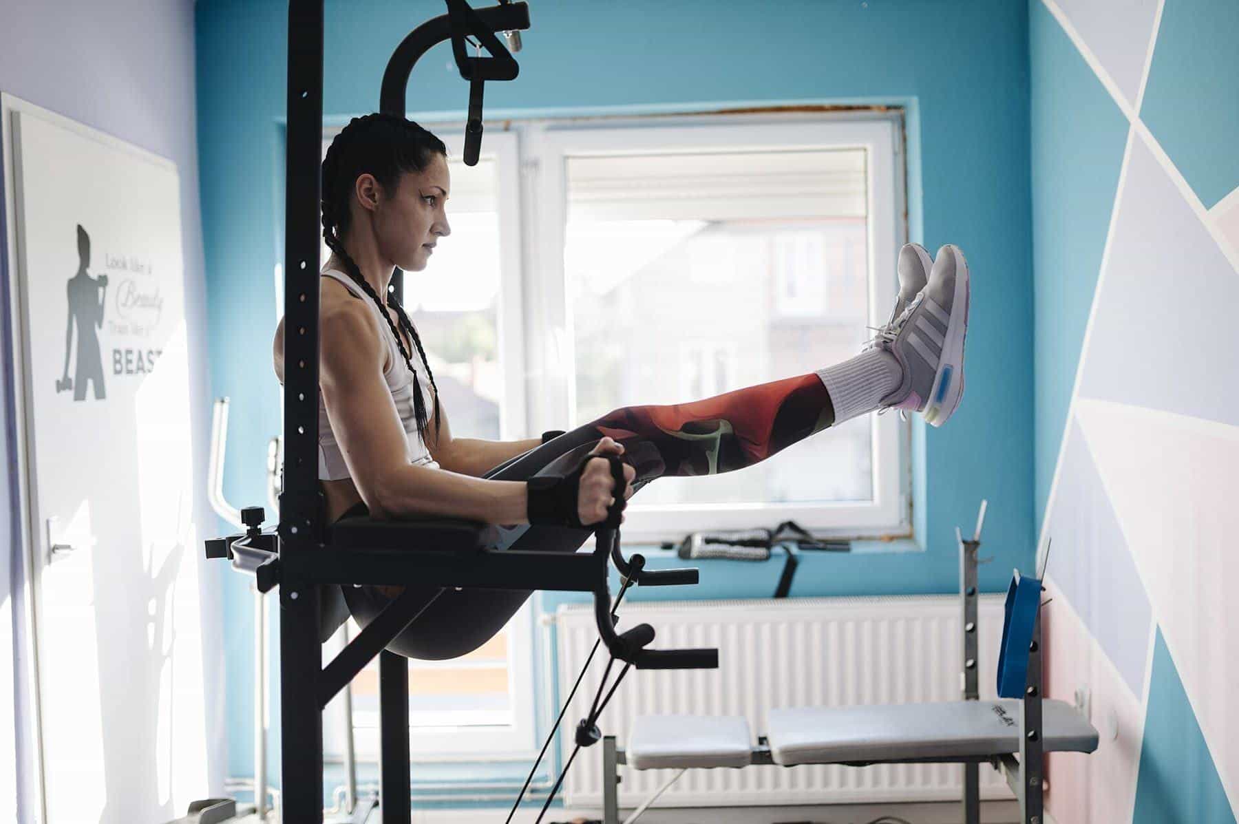 A woman doing knee raises on a dip station beside a bright window at home