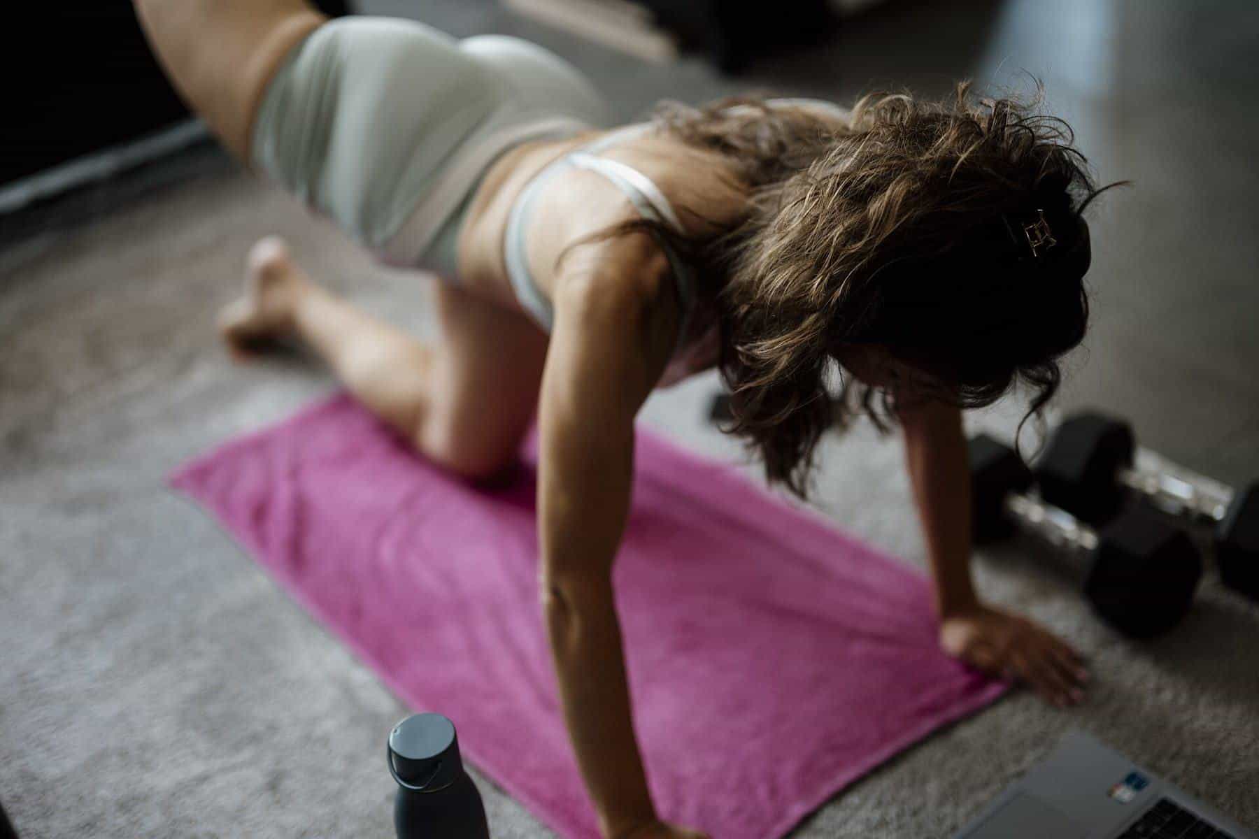 A woman stretching in a lunge on a pink mat at home with dumbbells beside her