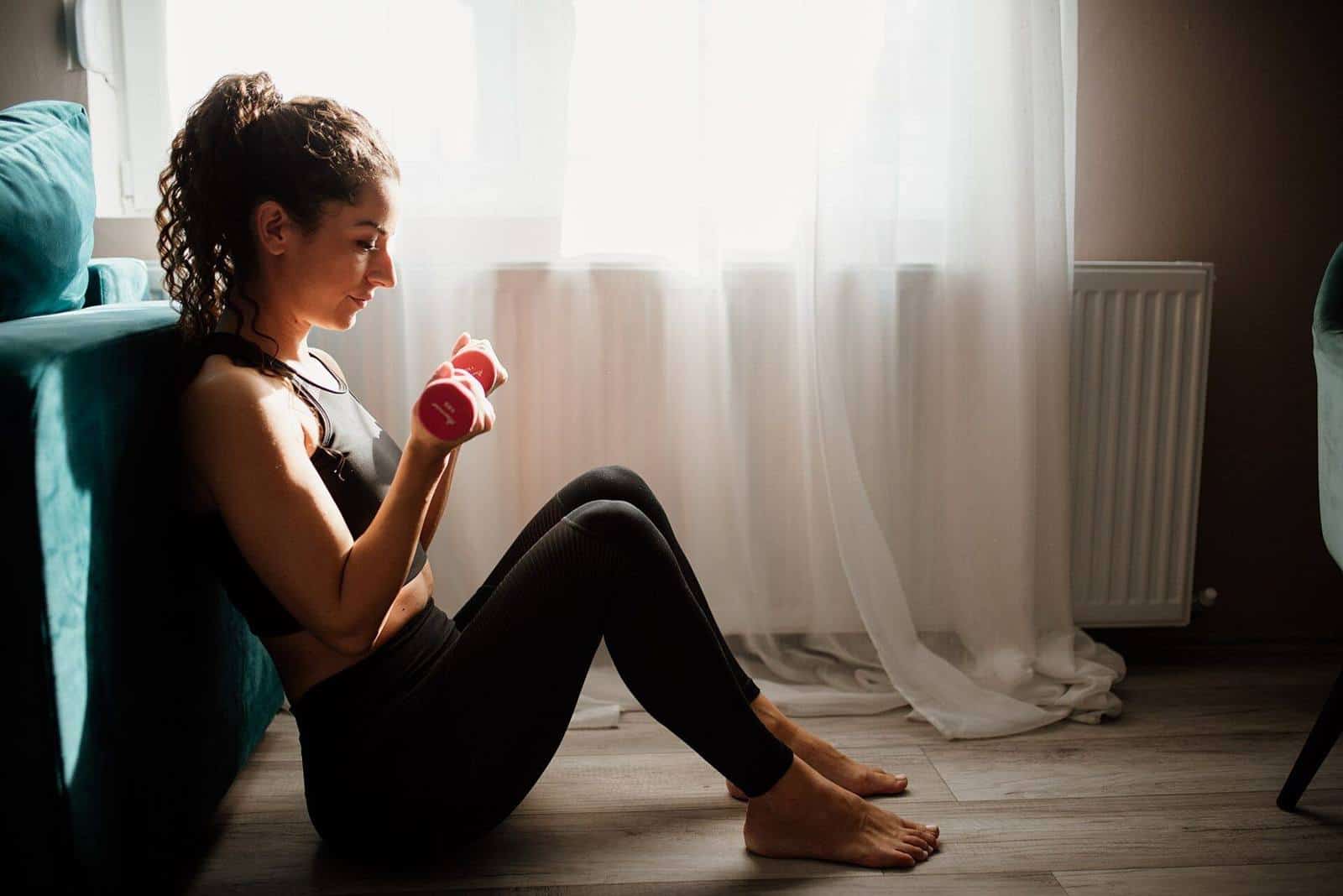 A woman sitting on the floor by a window doing a curl with a pink dumbbell