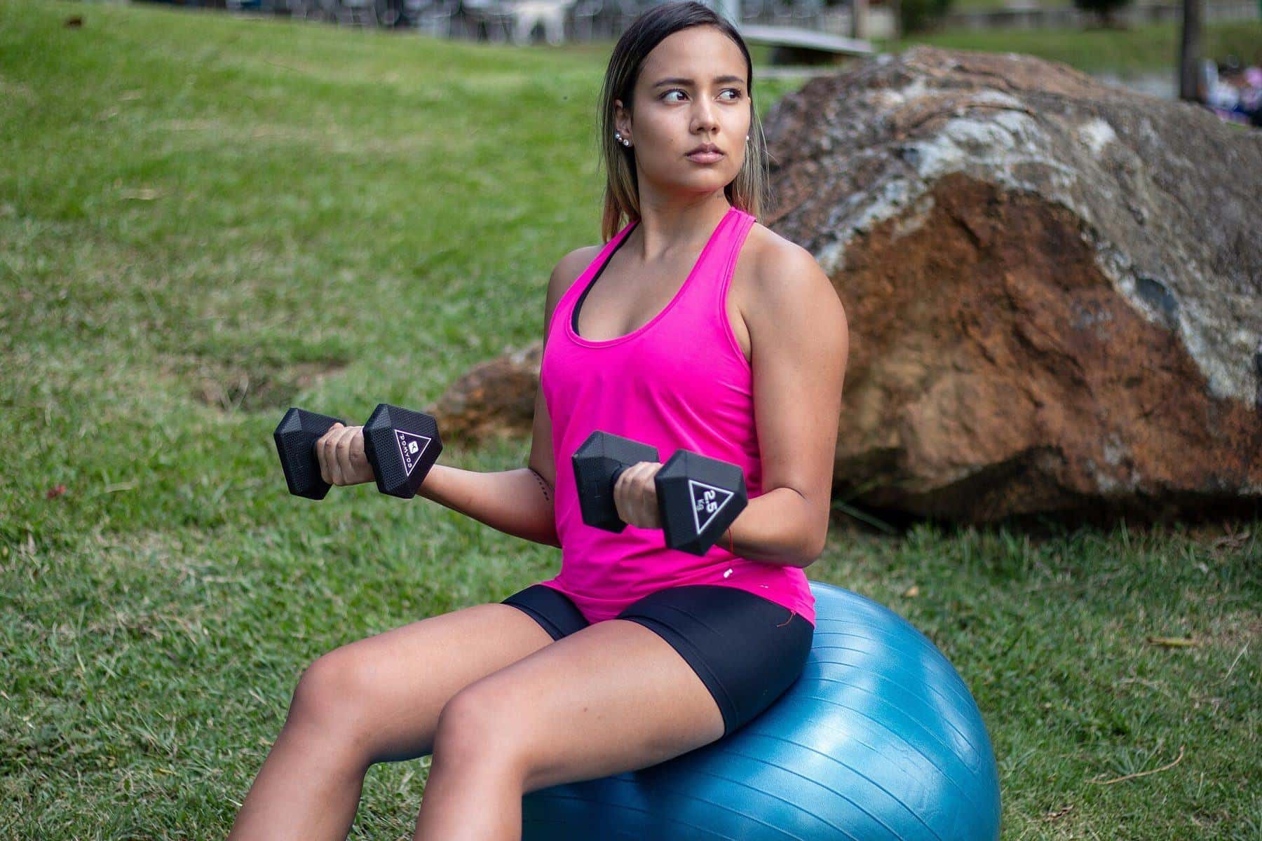 A woman doing bicep curls with dumbbells while sitting on a stability ball in a park