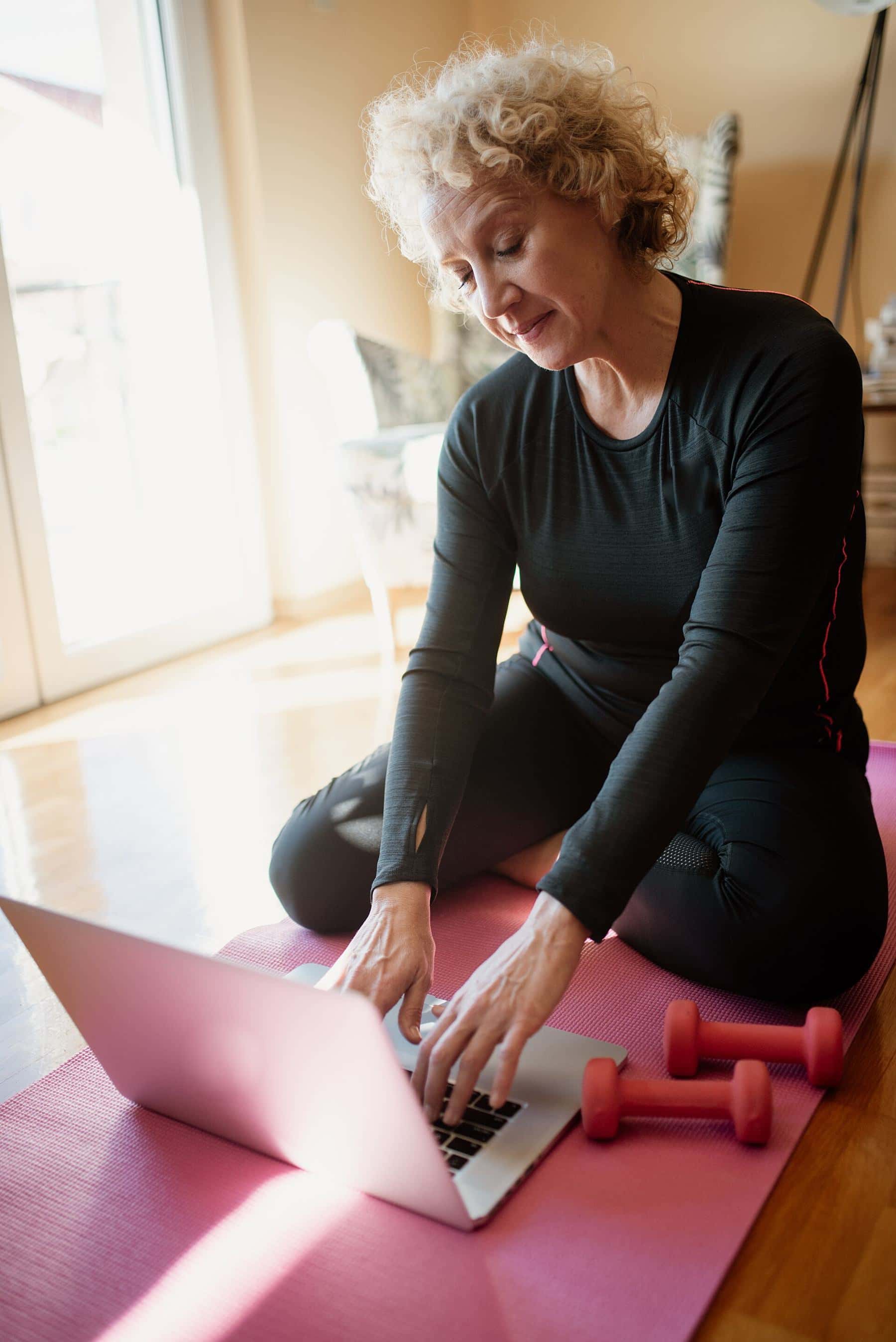 An older woman sitting on a pink mat with dumbbells, following a workout on a laptop