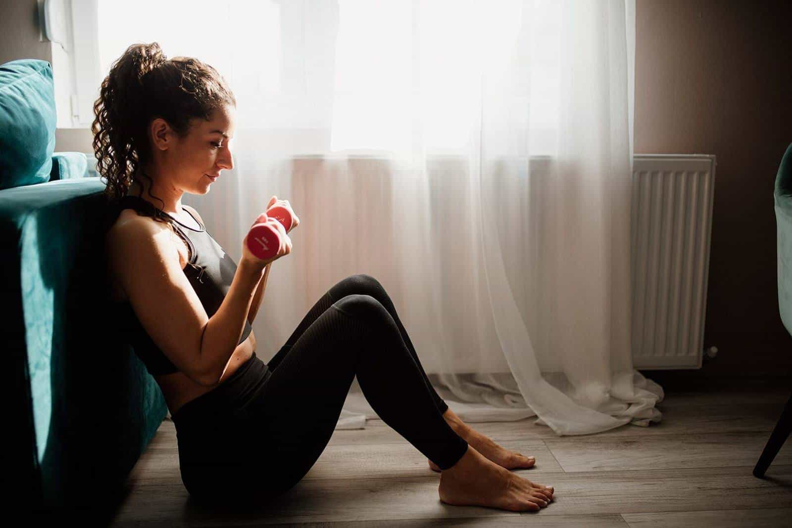 A woman sitting on the floor by a window doing a curl with a pink dumbbell