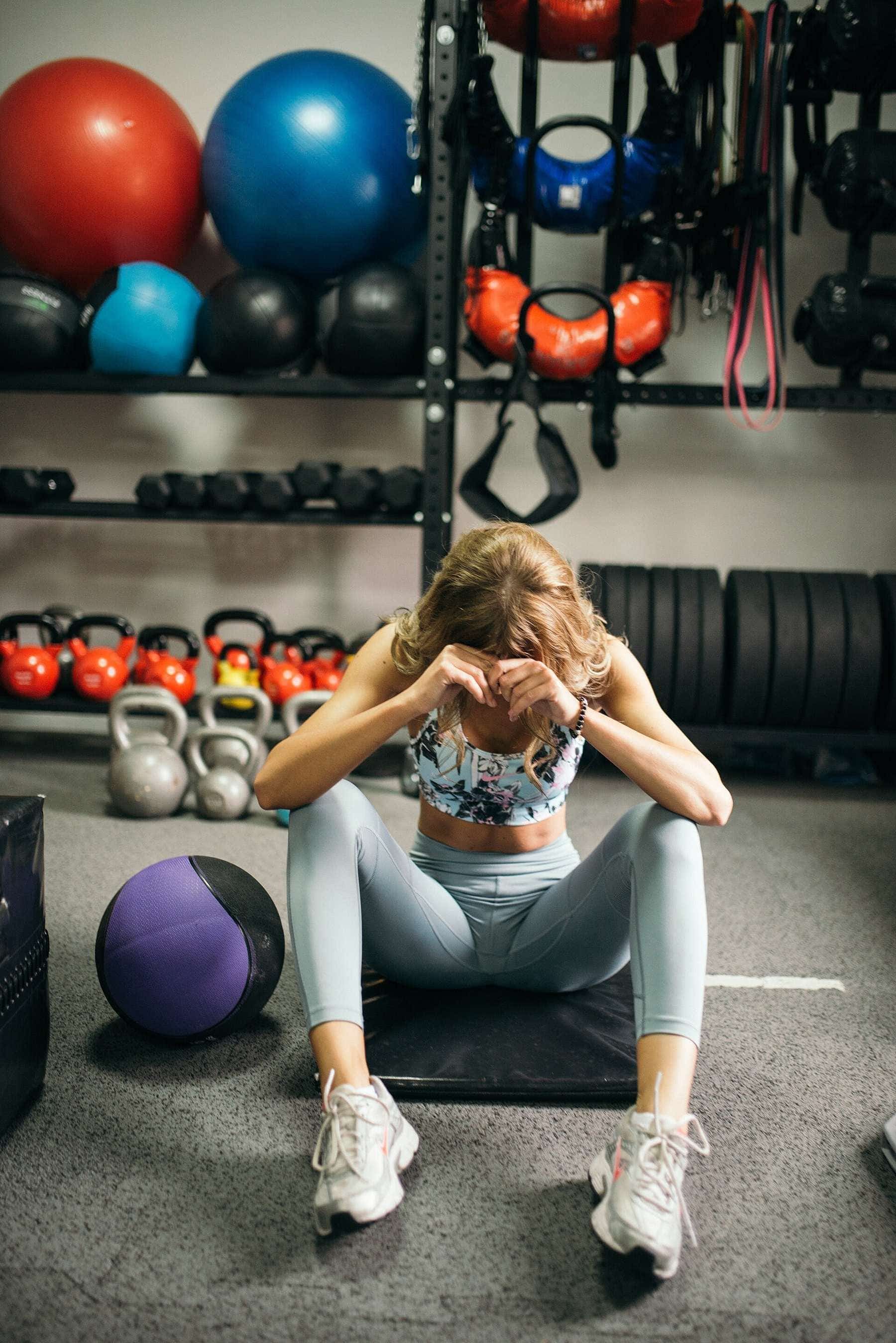 A woman sitting on a gym floor with her head in her hands after a workout