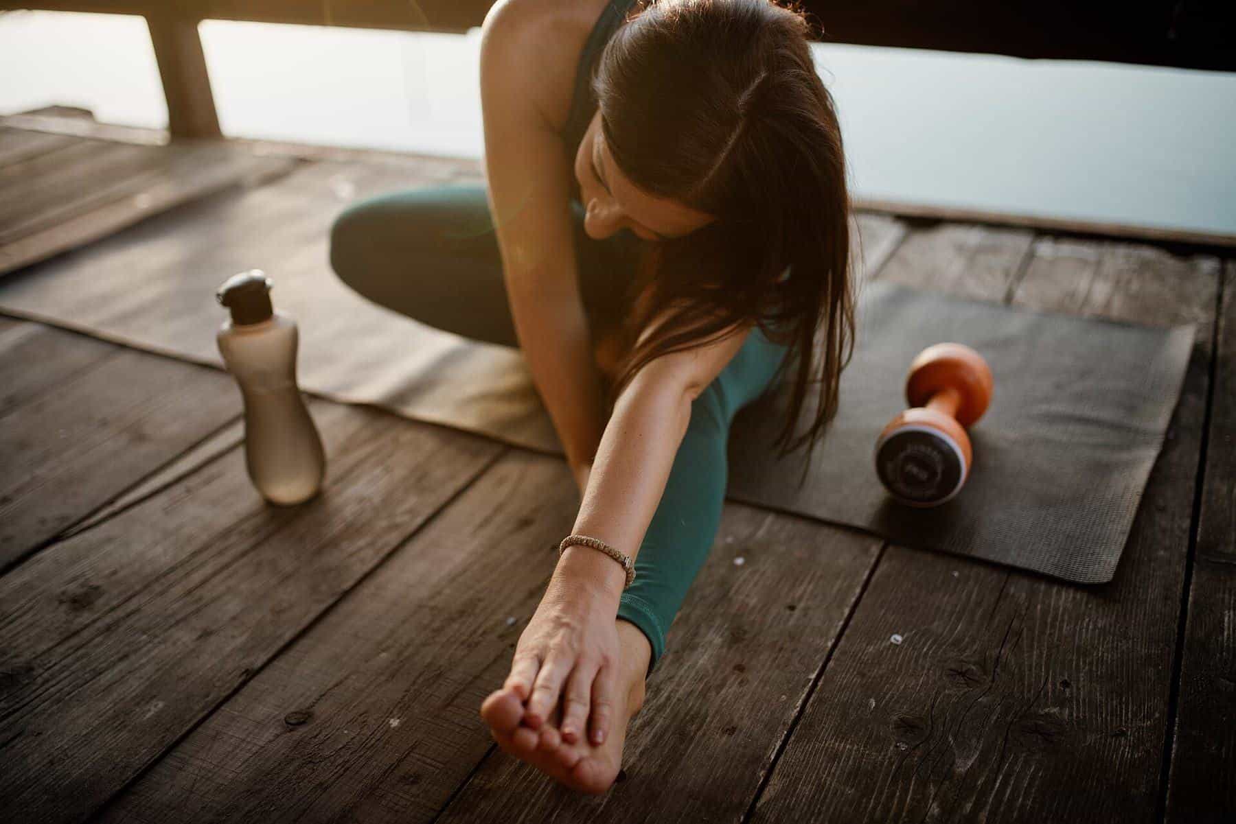 A woman stretching on a mat on a timber floor beside a water bottle and dumbbell